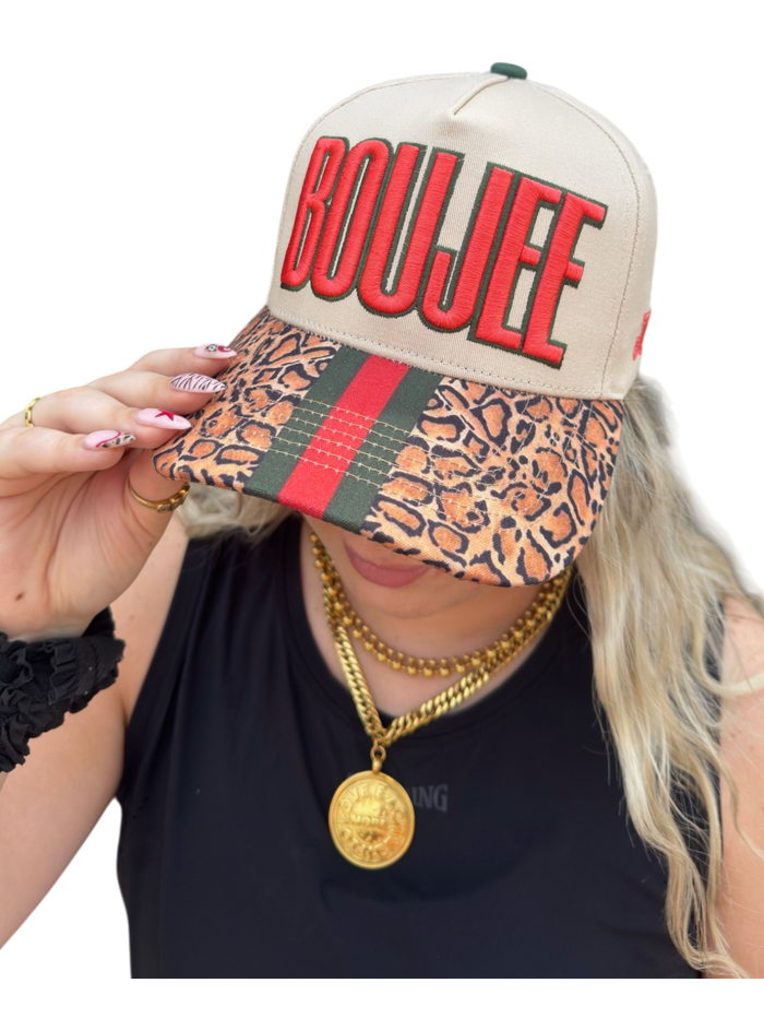 Person wearing a cap with 'BOUJEE' text and leopard print visor, holding it up.