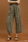 A model wearing olive green and beige checkered cropped wide-leg pants with a black top and dark green heeled sandals, set against a solid warm brown studio background.
