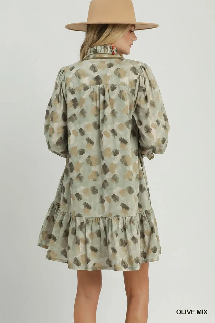 A woman shown from the back wearing a short olive green dress with a brown and beige abstract spotted pattern, long puff sleeves, and a ruffled hem. She is also wearing a wide-brimmed tan hat.