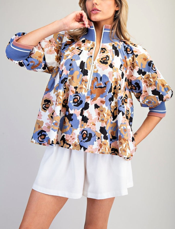 A woman models a short-sleeve floral print jacket with blue and pink striped ribbed cuffs and collar, paired with white shorts against a light gray studio background.