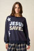 A woman wearing a navy blue crewneck sweatshirt with 'JESUS SAVES' printed in white and glittery gold letters, paired with navy plaid pants, against a solid beige background.