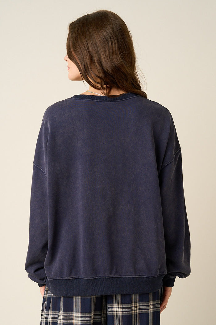 A woman viewed from behind wearing an oversized dark navy sweatshirt and plaid pants against a plain light beige studio background.