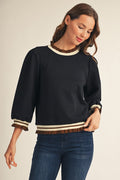 A woman wearing a black long-sleeve knit sweater with cream and brown striped ruffle trim at the crew neck, cuffs, and hem, paired with blue jeans against a light beige studio background.