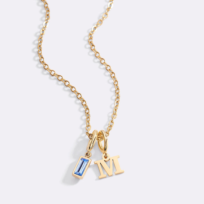Gold necklace with a blue gemstone pendant and 'M' letter charm on a light gray background