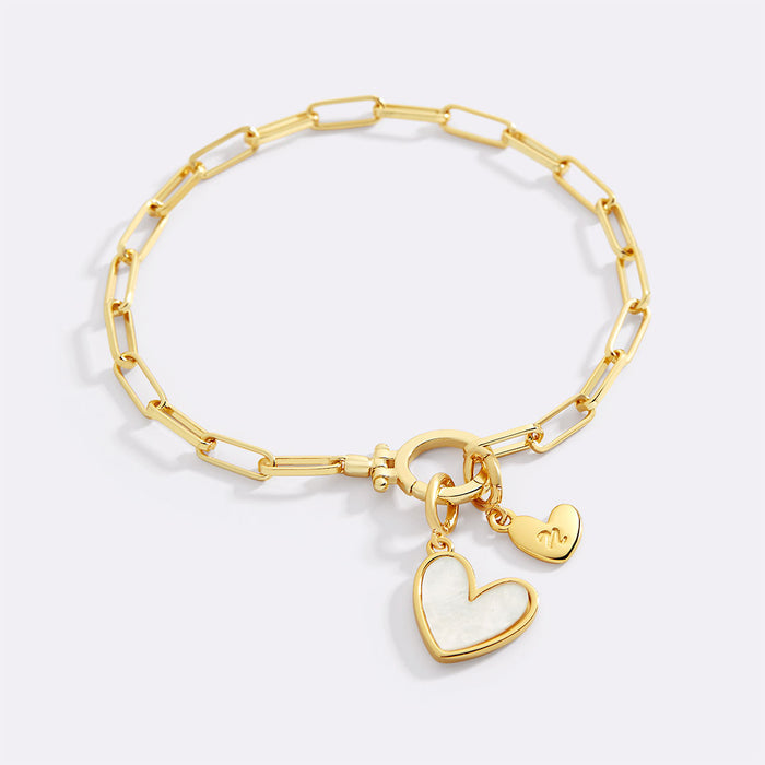 A gold bracelet with a heart-shaped charm and a customizable initial charm made from mother of pearl.