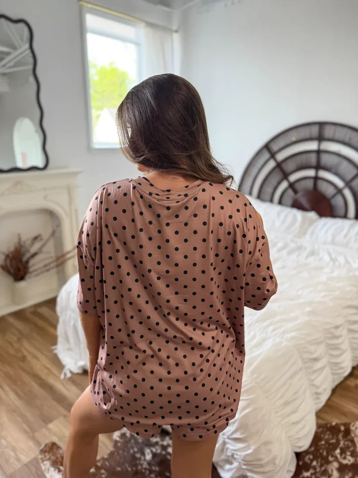 A woman viewed from behind wearing an oversized short-sleeve top and matching shorts in a dusty rose pink color with black polka dots, standing in a bedroom with a bed and fireplace in the background.